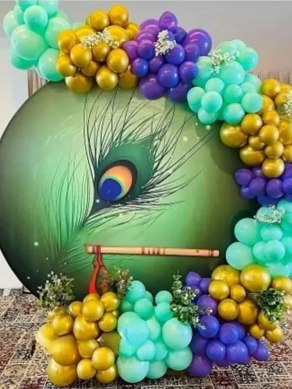 A beautiful and unique Janmashtami or Krishna-themed decoration. This setup features a circular backdrop with a large peacock feather design, surrounded by a balloon arch in vibrant shades of gold, teal, and purple.