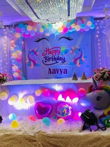 A magical unicorn-themed birthday stage. The backdrop features unicorn illustrations and is framed by pastel rainbow balloons, with a light-up unicorn prop and fairy lights.