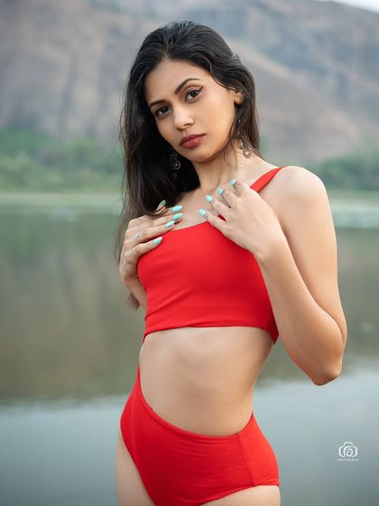 A close-up portrait of a model in a red swimsuit, with the calm lake and hills in the background. The focus is on her direct gaze and the vibrant color of her outfit.