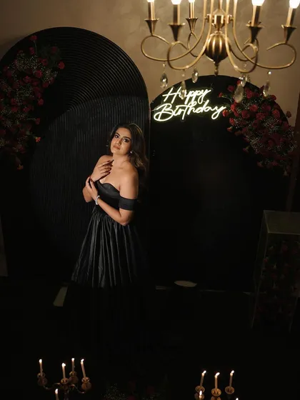 A chic and moody birthday photoshoot setting for an adult celebration. The decor features dramatic black arched backdrops, a stylish "Happy Birthday" neon sign, and romantic lighting from candelabras and a classic chandelier, proving birthdays are for grown-ups too.