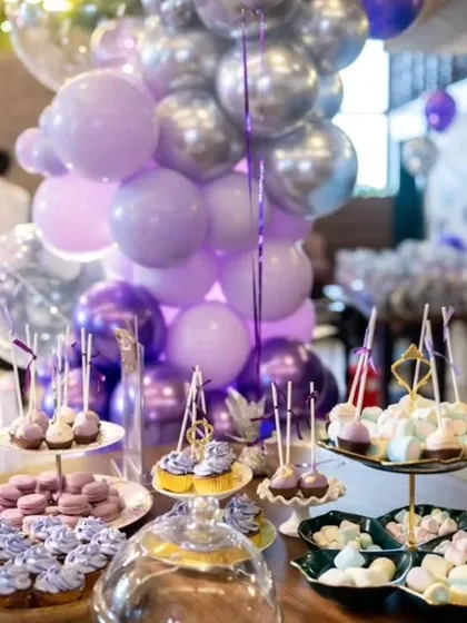 A beautifully styled dessert table with treats that match the party's purple color scheme. The display includes macarons, cupcakes, and cake pops, all arranged under a balloon centerpiece.