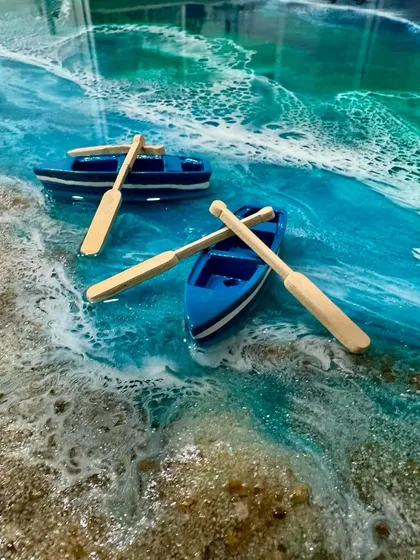 A close-up detail of a resin ocean art piece, showing miniature boats floating on realistic-looking water with sandy shores.