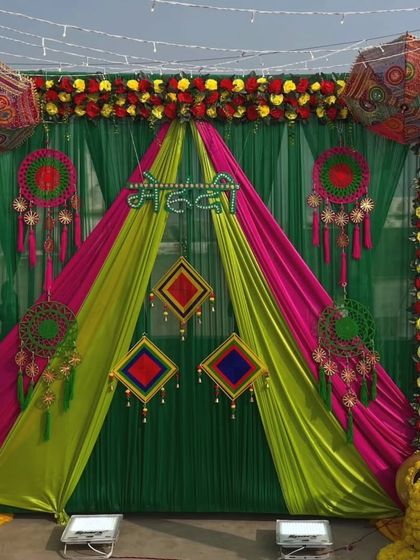 A vibrant Mehandi stage with a mix of green, pink, and yellow drapes. The decor is accented with Rajasthani umbrellas, floral borders, and traditional hangings.