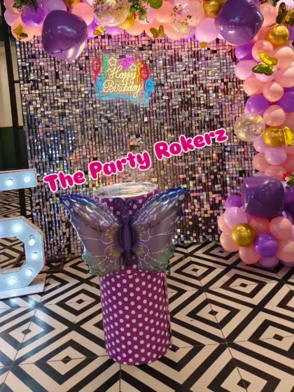 A full view of the purple butterfly theme, showing the arrangement of the balloon arch, the light-up number '5', and the butterfly-wing cake stand in front of a silver sequin wall.