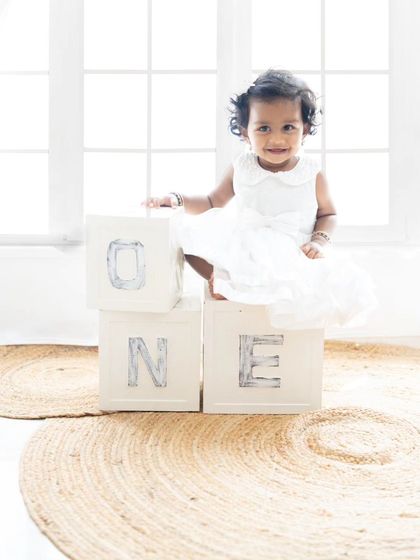 A beautiful, light-filled portrait of a baby girl celebrating her first birthday, sitting atop "ONE" blocks in a simple, elegant white dress.