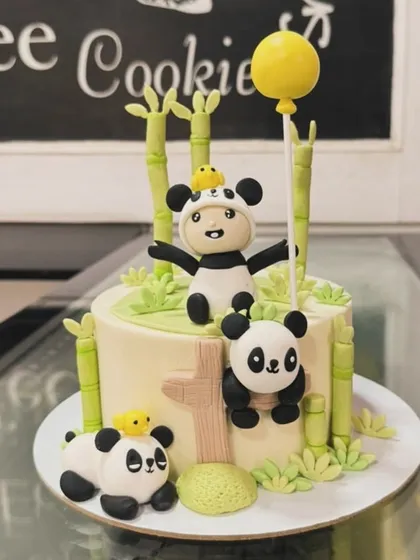 An adorable panda-themed cake for a first birthday. The design includes multiple cute panda figures, bamboo stalks, and a child in a panda costume.
