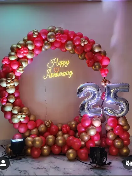 A vibrant circular balloon arch for a 25th anniversary, using a striking combination of red and gold balloons. The setup is completed with large silver foil numbers and a central "Happy Anniversary" neon sign.