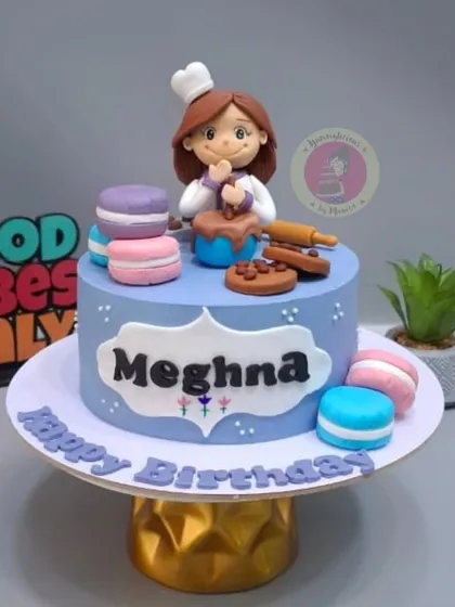 A cake for a fellow baker named Meghna, featuring a cute figurine of her in a chef's hat, mixing a bowl of batter.