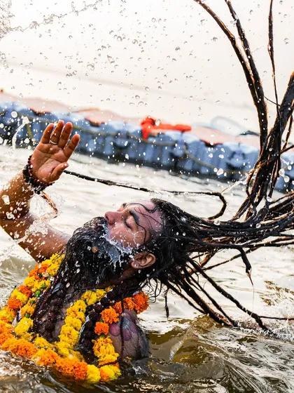 A Naga sadhu with long dreadlocks takes a holy dip in the Ganga, water splashing around him in a moment of spiritual ecstasy.