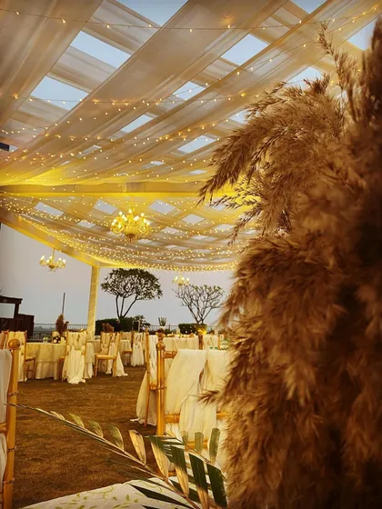 An elegant daytime setup with a boho touch. The tented ceiling with drapes and chandeliers is complemented by large arrangements of pampas grass, blending classic and rustic styles.