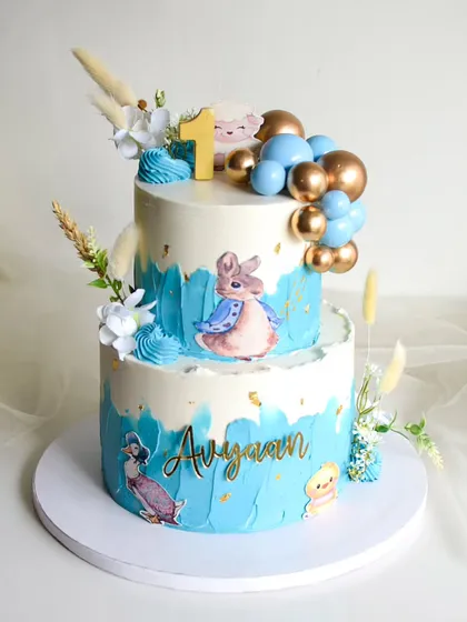 This Peter Rabbit themed cake was for a first birthday celebration. The two-tiered design has a blue watercolor effect and is decorated with edible character prints, gold and blue spheres, and dried grasses.