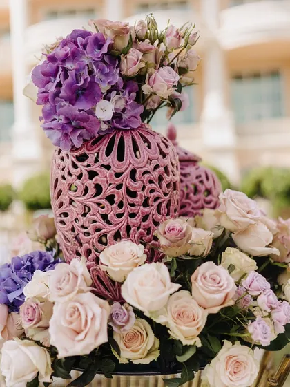 A close-up of a floral centerpiece, combining soft pink roses with vibrant purple hydrangeas in an ornate, carved vase.