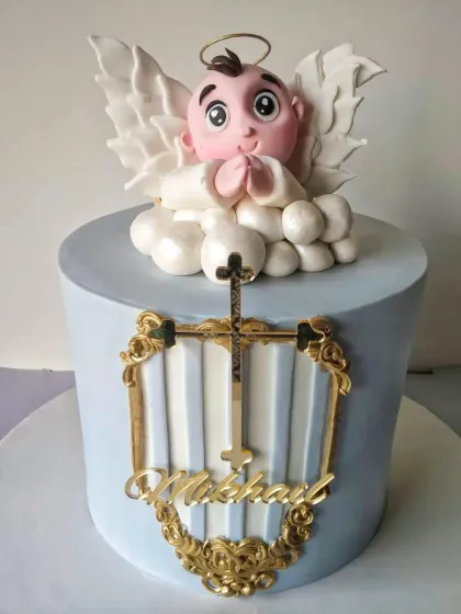 A beautiful blue and gold baptism cake. This design features an angel topper, a golden cross, and an ornate, shield-like plaque with the baby's name. The combination of pastel blue and rich gold creates a regal and reverent feel.