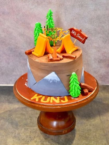 A fun camping-themed birthday cake for an outdoor lover. The cake is decorated with fondant tents, a campfire, pine trees, and a humorous "Bas 5 min!" sign.
