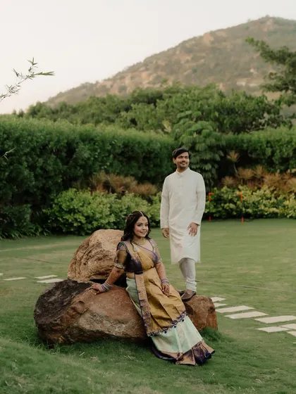 A relaxed portrait of a couple in a lush green estate. The bride is seated on a rock while the groom stands beside her, creating a natural and beautifully composed scene.