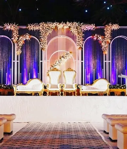 This reception stage is designed with modern arches, dramatic blue uplighting, and elegant white floral arrangements. The seating includes high-back throne chairs and a chaise lounge in white and gold, perfect for a contemporary royal theme.