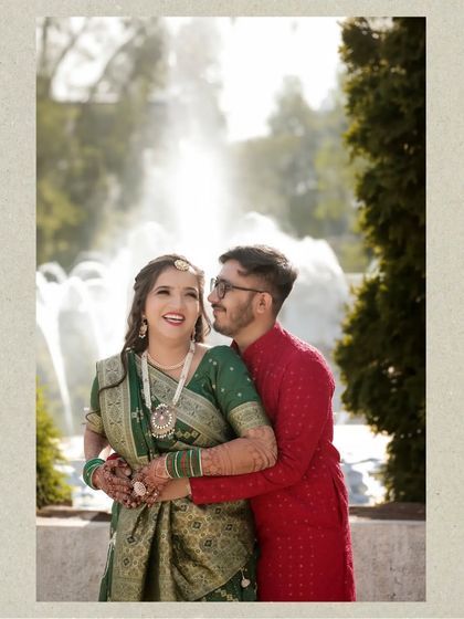 A joyful moment captured in front of a sunlit fountain. The groom embraces the bride from behind as she smiles, creating a warm and happy portrait in a lovely park setting.