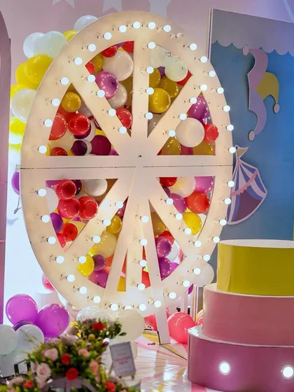 A lit Ferris wheel prop filled with colorful balloons serves as a stunning centerpiece for this carnival stage. The tiered cake stand in front is ready for the main event.