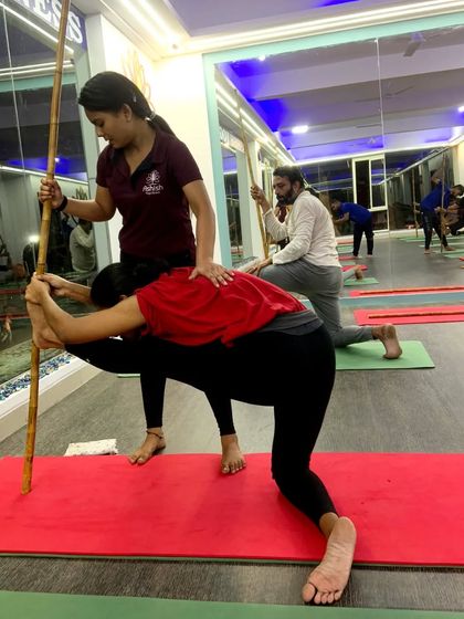 An instructor uses a yoga stick to help a student deepen her hamstring stretch. This prop-assisted adjustment ensures correct alignment and a more effective stretch.