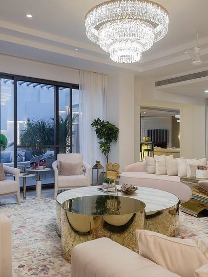 A wider view of the blush-toned living room, showing the full seating arrangement and the grand tiered chandelier. Large glass doors connect the space to an outdoor patio, enhancing the feeling of light and airiness.