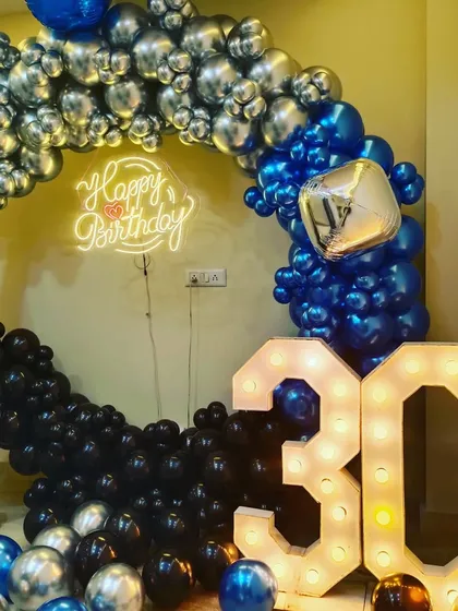 Celebrating a 30th birthday in style. This modern setup uses a ring of chrome silver, blue, and black balloons, a large light-up number "30", and a neon sign for a cool, party-ready photo corner.