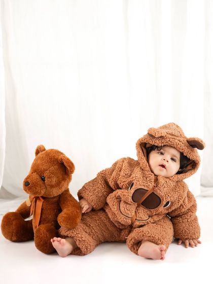 A seven-month-old baby in a fluffy teddy bear costume lies next to his teddy bear friend. This is when they can often sit up, a major milestone to celebrate.