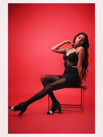 A full-length shot of a model in a glamorous black outfit against a red background. The pose is strong and confident, creating a striking fashion image.