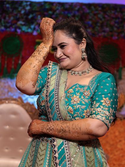 A candid shot of a bride smiling, her hand gently touching her head, showcasing the beautiful mehandi on her arm.