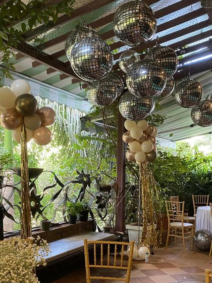 An outdoor patio decorated for a 60th birthday party. The ceiling is adorned with sparkling disco ball balloons, while balloon columns in neutral tones add a festive touch to the lush green surroundings.