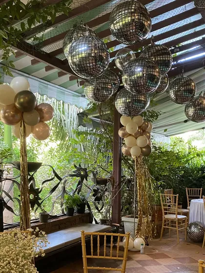 An outdoor patio decorated for a 60th birthday party. The ceiling is adorned with sparkling disco ball balloons, while balloon columns in neutral tones add a festive touch to the lush green surroundings.