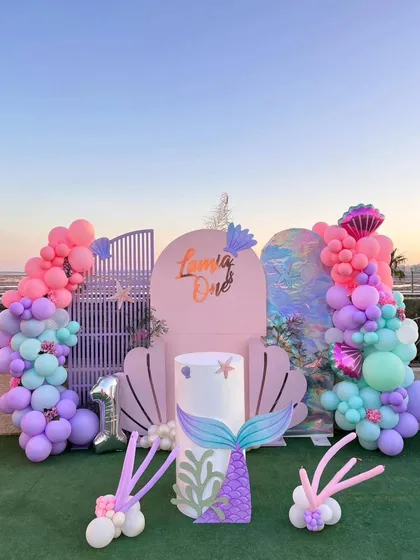 Dive into an under-the-sea adventure with this mermaid-themed first birthday party. The setup features a large seashell photo booth, mermaid tail accents, and a vibrant balloon garland in shades of pink, purple, and teal, creating a magical ocean kingdom for your celebration.
