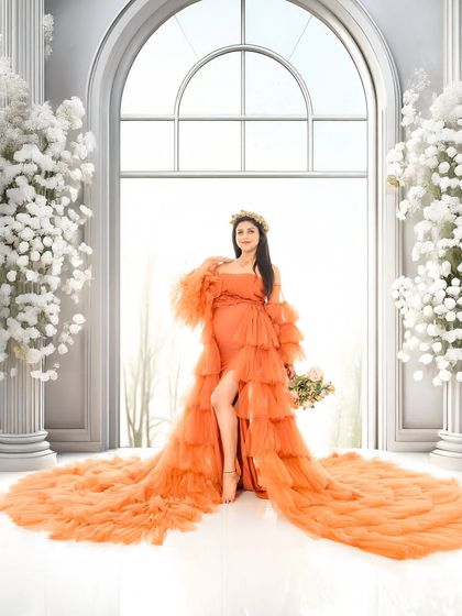 A powerful and confident solo portrait of the mom-to-be in a vibrant orange ruffled gown. The grand white arches in the background add a sense of scale and drama.