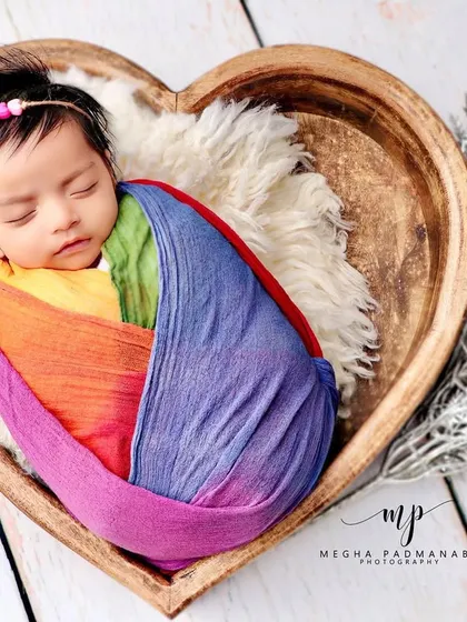 A baby wrapped in a vibrant rainbow swaddle, nestled in a heart-shaped wooden bowl.