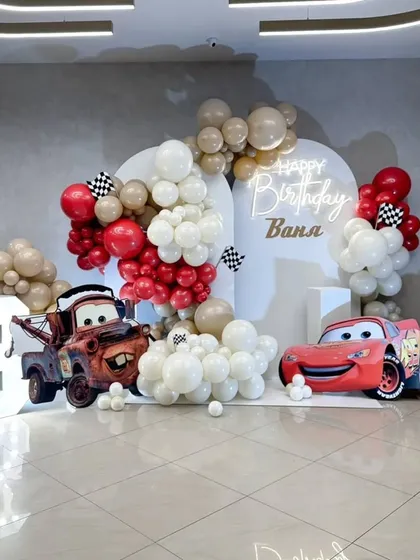 A high-energy Disney Cars theme for a 3rd birthday. This setup includes character cutouts of Lightning McQueen and Mater, a custom backdrop with a neon name sign, and balloon arrangements in a clean red, white, and beige color scheme.