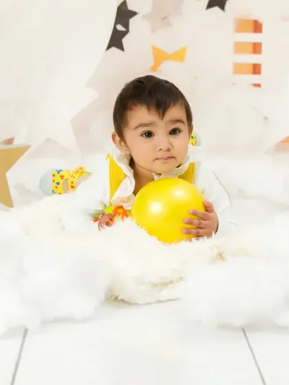 A quiet moment during a cake smash session, where the baby lies among soft, cloud-like props, holding a yellow ball.