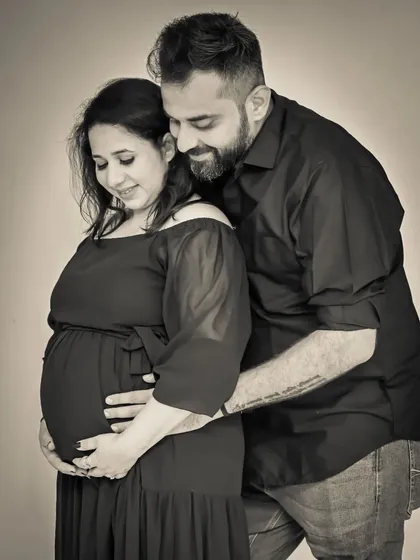 A tender black and white studio portrait. The dad-to-be embraces his partner from behind, both looking down at the bump with love.