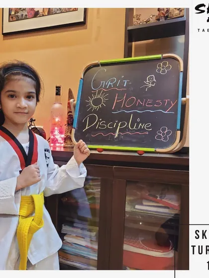 Grit, Honesty, Discipline. A young student stands proudly beside a board displaying the core values we instill in our classes, ready to practice her Taekwondo forms.