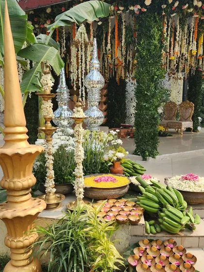 A South Indian wedding setup rich in tradition. The decor features floral gajras, banana bunches, and diyas on banana leaves, with tall golden and silver lamps adding to the sacred ambiance.