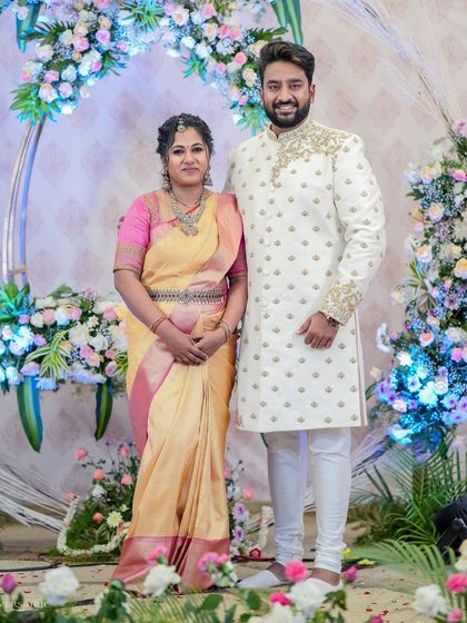 A couple posing against a floral backdrop. His white embroidered sherwani and her yellow and pink saree are a perfect example of our bespoke couple's wear.