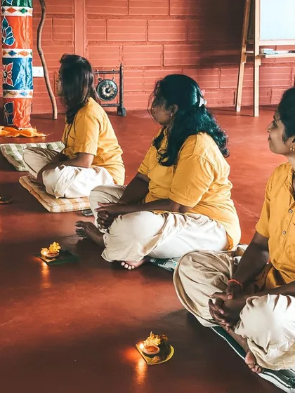 Three students sit in quiet contemplation during a ceremony, with small lamps (diyas) placed before them. This practice of offering light is a beautiful part of our rituals.