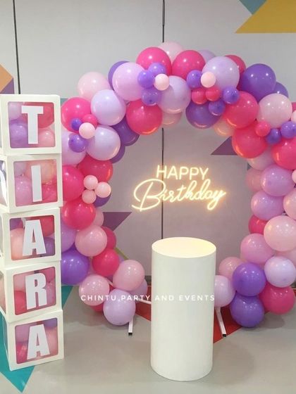 A vibrant balloon ring in shades of pink and purple for a birthday. The setup is personalized with transparent boxes spelling the name 'Tiara' and includes a pedestal for the cake.
