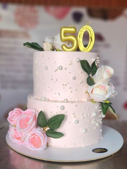 A lovely two-tier cake for a 50th anniversary celebration. The soft pink frosting is dotted with edible pearls and decorated with beautiful artificial roses, topped with golden '50' candles.