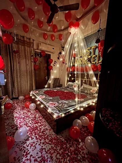 Here is a full view of a romantic room setup, showing the floor completely covered in red rose petals. This design includes a fairy light canopy over the bed, 'Just Married' balloons, and dozens of red and white balloons on the ceiling.