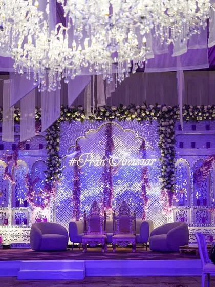 A breathtaking stage for an engagement ceremony, bathed in lavender light. The backdrop features intricate jaali work, floral adornments, and a custom neon hashtag, all under a canopy of crystal chandeliers.
