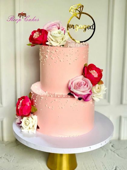 A lovely two-tier engagement cake in a soft pink hue. The cake is decorated with pearl-like sprinkles and adorned with beautiful red, pink, and white roses for a classic, romantic feel.
