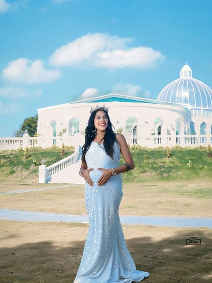 A majestic outdoor portrait of a mother-to-be in a sparkling white gown and crown. The grand architectural background adds a touch of royal elegance to the maternity shoot.
