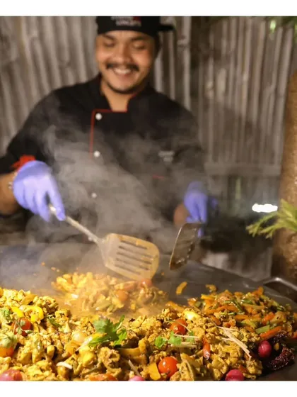 A happy chef smiles as he prepares a large batch of scrambled eggs or a similar dish on a tawa. The joy our team takes in their craft is evident and contributes to the positive atmosphere of our events.