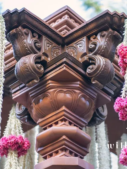 A detail of the intricately carved wooden pillar of the wedding mandap, adorned with fresh flower garlands.