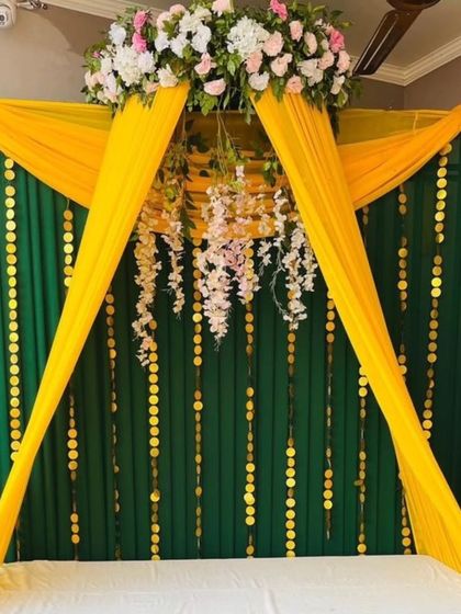A stylish Mehandi seating with a canopy created from yellow drapes and a floral centerpiece. The backdrop of green fabric with golden disc hangings adds a touch of glamour.