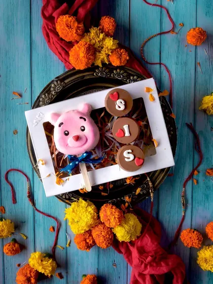 Rakhi is a special celebration of the bond between siblings. This adorable hamper for a sister includes a cute pink teddy bear cakesicle and Oreo-filled chocolates that spell out "SIS," all beautifully presented in a festive box.